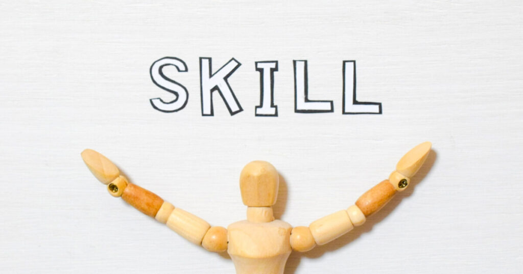 「SKILL」の文字の下で両腕を広げて立つ木製デッサン人形。スキルアップや能力開発のメタファーとして、成長と可能性を表現している。